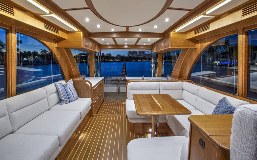 The teak-trimmed saloon of a motor yacht at dusk, with upholstered seating and the waterfront visible through the windows