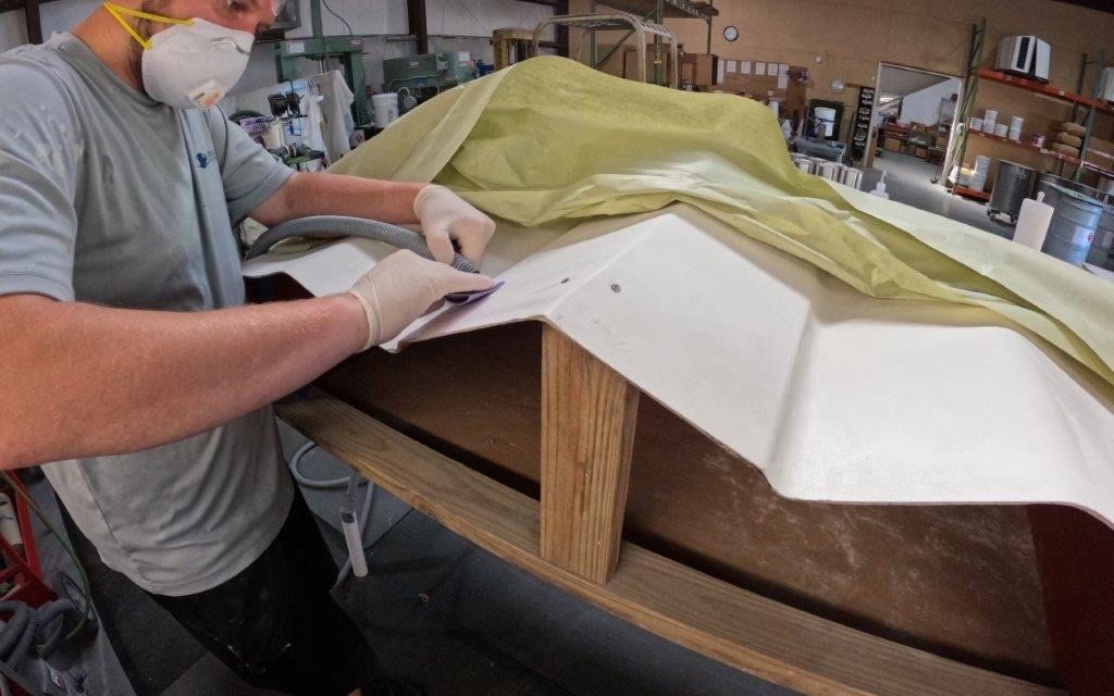 A technician in a respirator and gloves sanding a fiberglass hull section in the repair shop